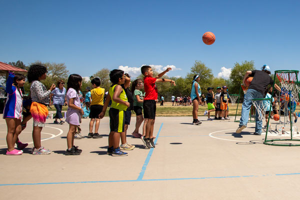 Line of children wait turns whil a basketball soars towards its hoop.